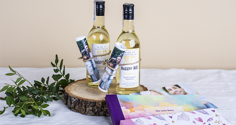 Due piccole bottiglie di vino bianco decorate con le foto di una coppia innamorata, arrotolate e legate con uno spago di iuta. Accanto si può vedere un rametto verde e alcuni sharebook firmati Colorland.