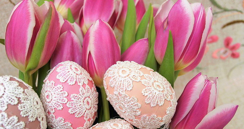 Velikonoční vejce zdobené krajkou, v pozadí růžové tulipány.