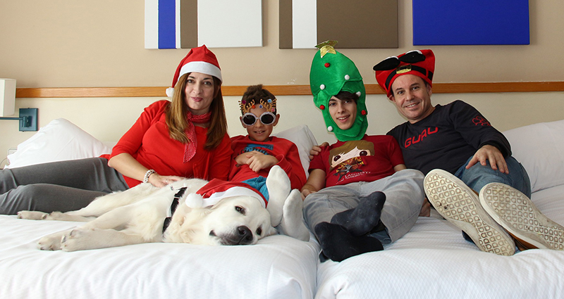 Familiebeeld met vier personen en een hond. Ze liggen op een bed – iedereen in een kerstmuts. 