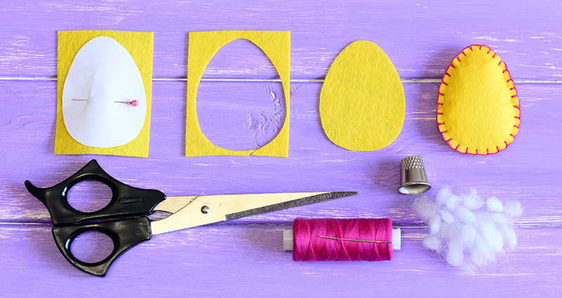 Des œufs  en feutre, des ciseaux, un fil, un dé à coudre et du coton sur une table violette en bois.