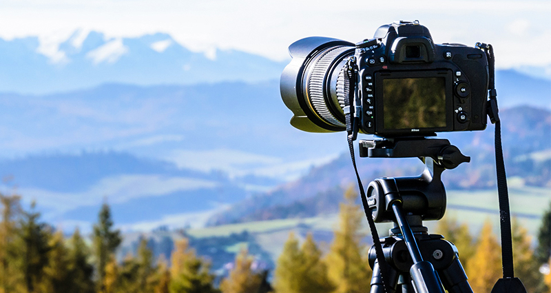 Focus on a camera mounted on a tripod, wood and mountains in the background