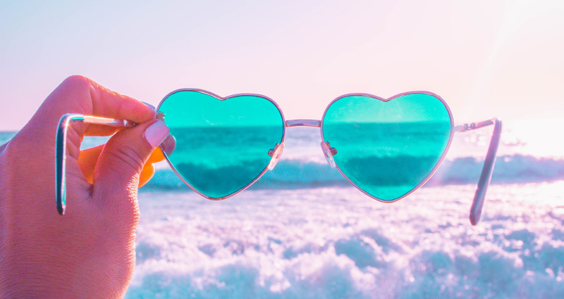 Focus on a woman’s hand holding heart shaped sunglasses, sea waves in the background. There is a pink filter which has been added to the photo.
