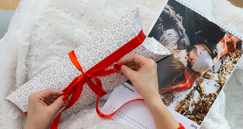 Detailní záběr na ruce ženy odpočívající pod dekou, která uvazuje červenou mašli na kalendářovém balení, vedle fotokalendář.