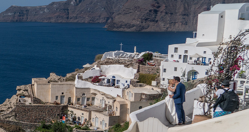 La foto di una coppia di sposi che si baciano su una delle terrazze di Santorini. Sulla foto presente anche il fotografo che scatta loro delle foto e il mare sullo sfondo.
