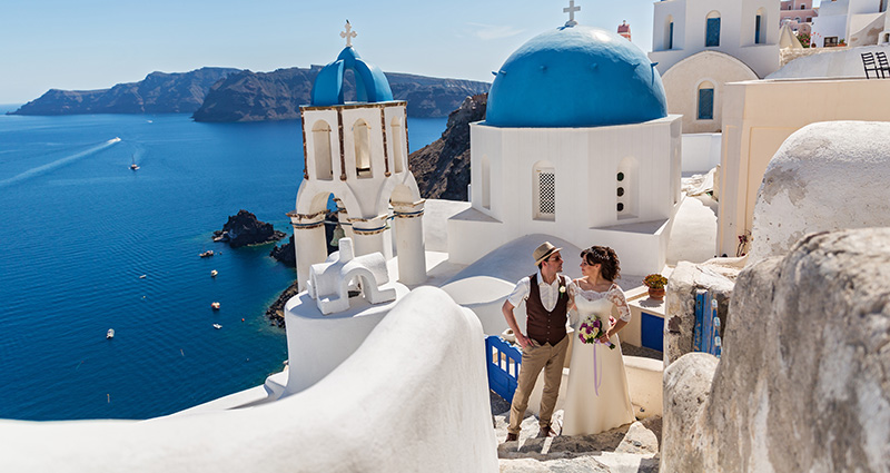 La foto raffigura una felice coppia di sposi a Santorini; sullo sfondo sono visibili le caratteristiche chiese con i tetti blu.