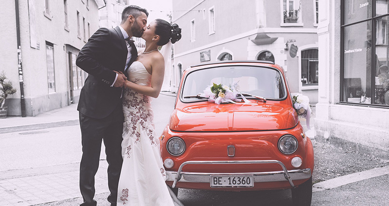 A photo of a newlywed couple kissing in an Italian alley. An old, red fiat near them.