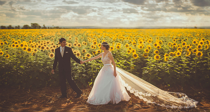 Ein Foto eines Brautpaares, das nahe bei einem blühenden Sonnenblumenfeld spazieren geht, im Hintergrund bewölkter Himmel.