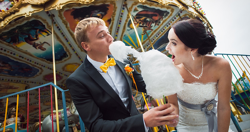 A photo of a newlywed couple eating cotton candy in an amuesement park. A carousel behind them.