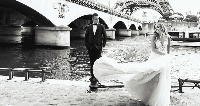 Čiernobiela fotografia nevesty a ženícha pri nábreží Seiny, v pozadí most a Eiffelova veža.
