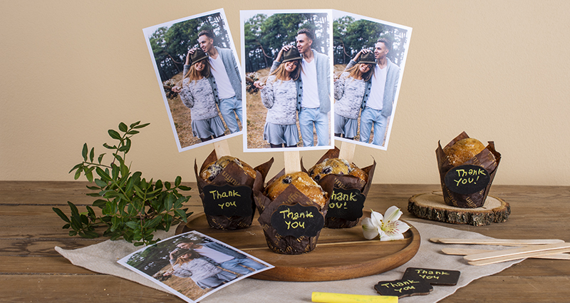 Rectangular photos glued to ice cream sticks and sticked into muffins with 'Thank you' caption on them. Muffins on a wooden tray placed on a light cloth and dark table, a print, green brunch, yellow  chalk and thank you cards around it.