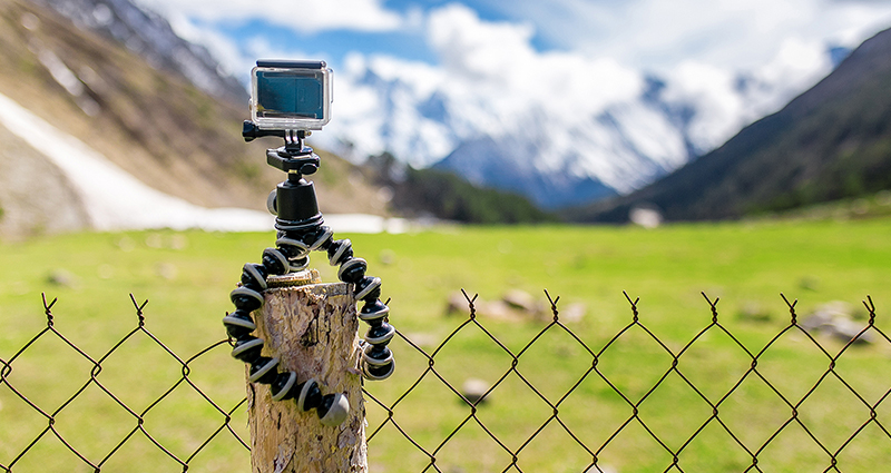GoPro-Kamera auf einem mobilen Stativ mit Gorillapod-Halterung an einem Holzpfosten, im Hintergrund ein Blick auf die Wiese, Berge und Himmel.