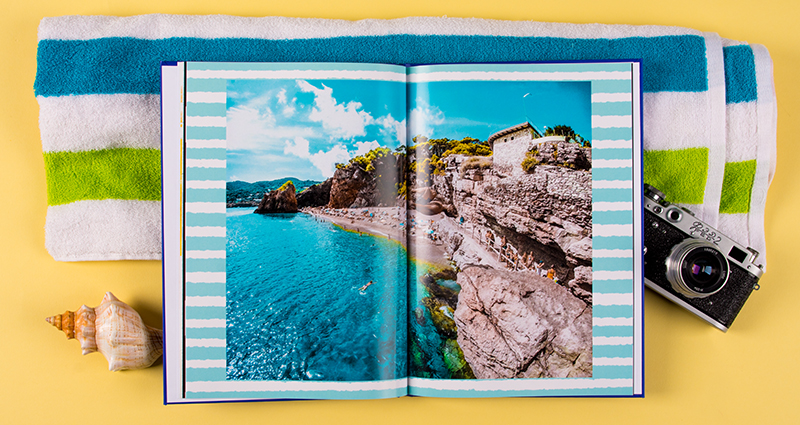 Holiday photobook with a picture of a rocky seashore, a blue and green striped towel in the background, next to a camera and a shell