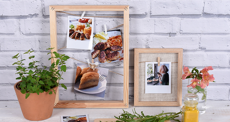 Insta photos and retro prints of food, some of them in a frame, others lying on the table next to potted herbs and a chopping board with cherry tomatoes