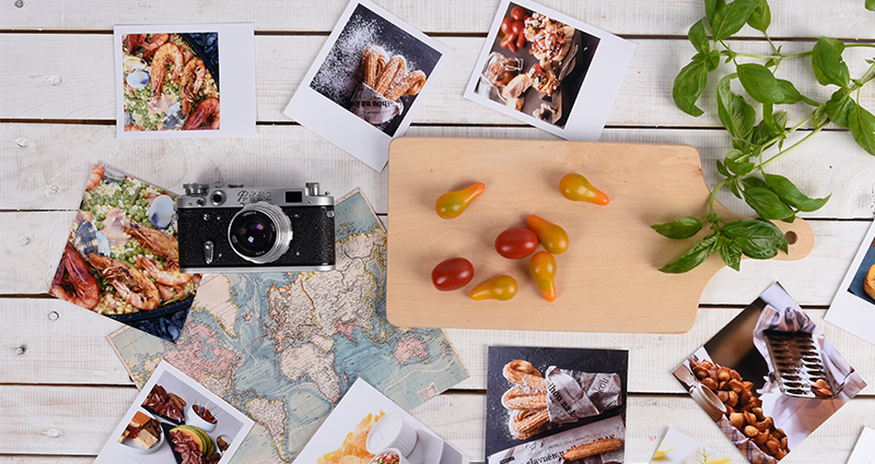 Insta fotos y revelados retro que representan la comida, un mapa del mundo en el fondo, sobre el mapa - una cámara, al lado – una tabla de cortar con tomates cherry y una ramita de albahaca.