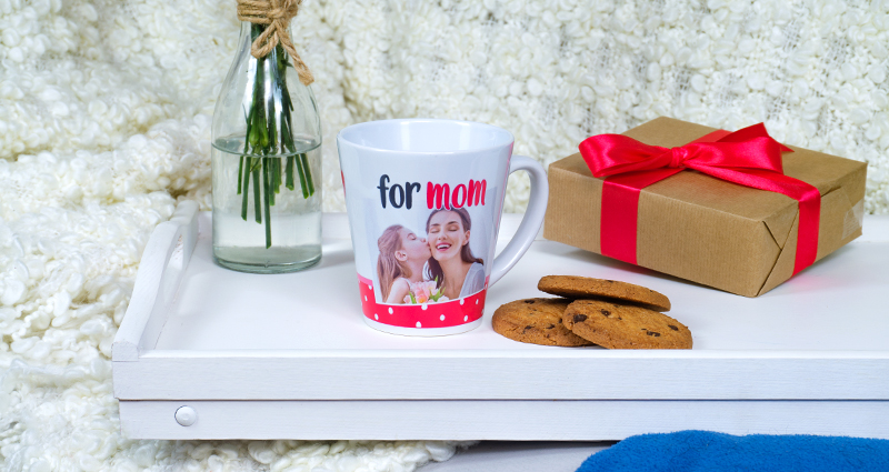 Un mug latte avec une photo de maman et sa fille et le texte „for mom” sur un plateau blanc,  à côté  une vase avec des fleurs et un cadeau emballé.