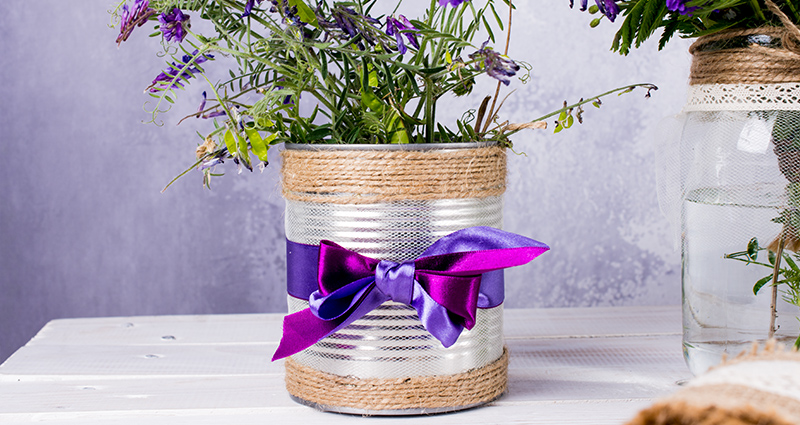 Flores silvestres en una lata decorada con una malla, una cuerda de yute y cintas. Al lado unas flores en una jarra.