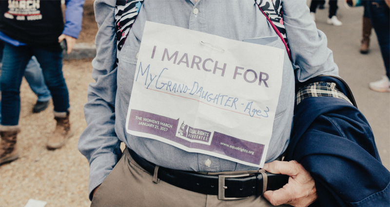 Old man holding a piece of paper with note on it: "I march for my granddaughter, age 9".