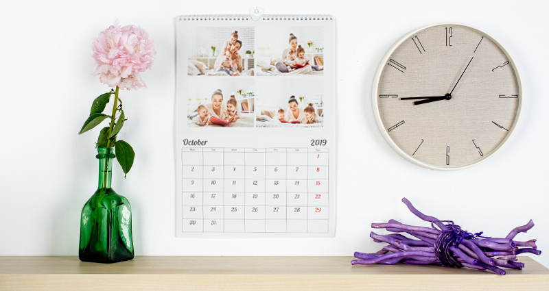 Photo calendar with 4 photos hanging on a wall next to a round clock; there is a shelf with a flower vase below it.