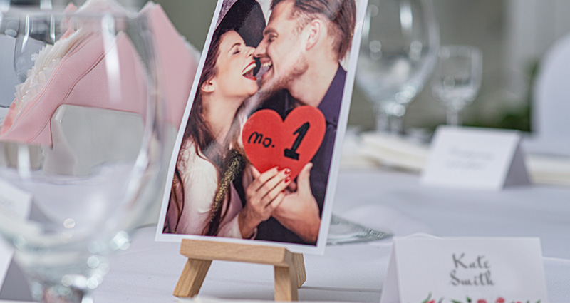 Fotografie zamilovaného páru držícího červené srdce s nápisem "No. 1 "na dřevěném mini stojanu, na stole pokrytý bílým ubrusem; kolem skleněné pohárea a jmenovky.