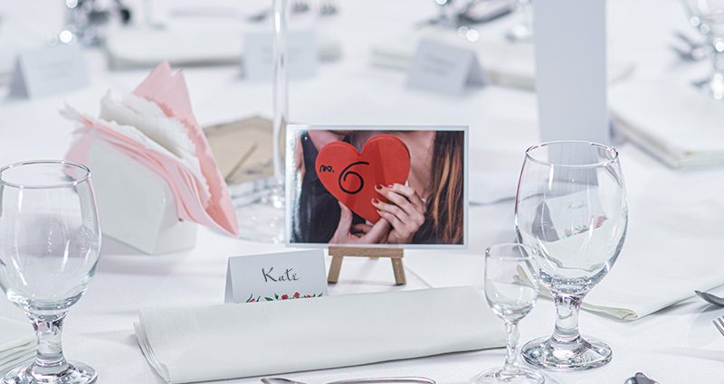 Fotografia zamilovaného páru držiaceho červené srdce s nápisom „No. 6“ na drevenom mini stojane, na stole pokrytý bielym obrusom; okolo sklenené pohárea a menovky.
