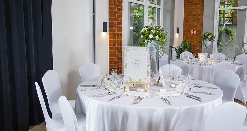 Ein Bild eines weiß verzierten runden Hochzeitstisches im Bankettsaal; auf dem Tisch Tafelservice, Blumenstrauß und pastellfarbene Fotoleinwand mit der Tischnummer und mit den Dankessprüchen für die Gäste.