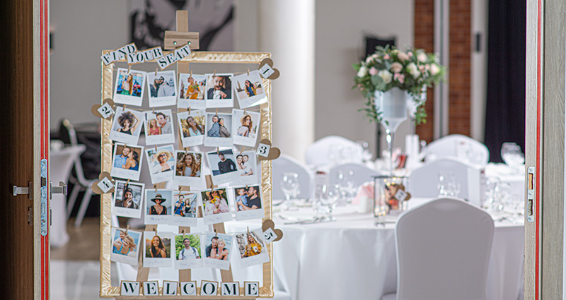 Un marco hecho a mano con fotos de invitados a la boda y los números de mesas. En la parte superior la inscripción "Encuentra tu asiento", en la parte inferior "Bienvenidos", en el fondo una mesa en el salón de banquetes. 