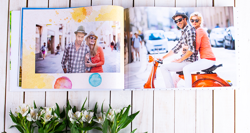 Photos of a couple in a pastel-coloured album on a white wooden background, flower decorations below.