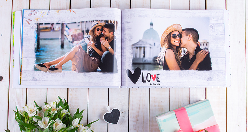    Įsimylėjusios poros nuotraukos albume, po viena iš nuotraukų tekstas: Love is in the air. Fotoknyga ant balto medinio fono, žemiau gėlių dekoracijos ir spalvinga dovanų dėžutė.