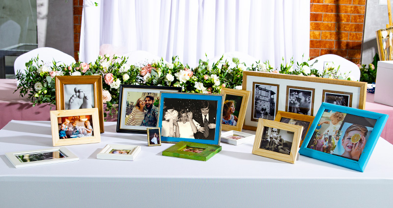 Oude huwelijksfoto's van de ouders van de bruid en bruidegom en jeugdfoto's van de pasgetrouwden geplaatst in kleurrijke lijsten op een witte tafel (geheugenhoek); op de achtergrond - een tafel versierd met bloemen en felle lantaarns aan beide zijden van de vloer.