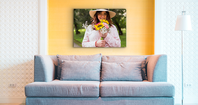 Nuotrauka pilkos sofos su geltona siena fone. Ant sienos didelis fotopaveikslas su nusišypsojusia moterimi su geltonai rožinės spalvos gėlių puokšte.