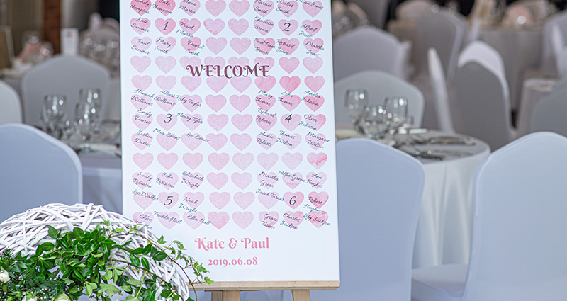 Lienzo con foto de una lista de invitados a la boda, hecho de una plantilla de acuarela con corazones, en un caballete. Con mesas blancas en el fondo y un adorno floral al lado. 