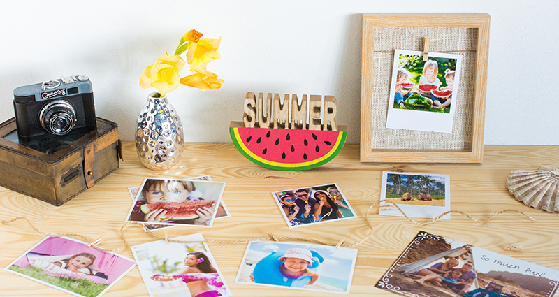 Rectangular prints, insta photos and retro print on the desk; a wooden photo frame, flowers in a flower vase, a camera and other accessories in the background