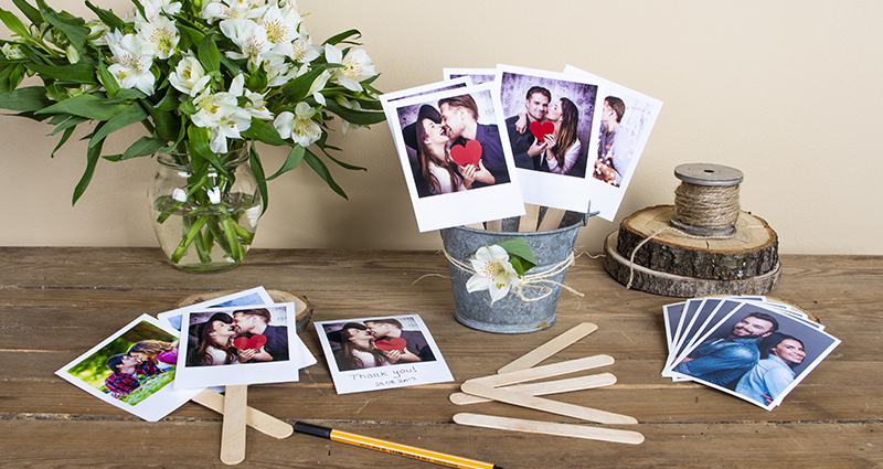 Retro prints on an ice cream sticks in a decorative pot, arranged photos, sticks and black marker placed next to it, white flower bouquet in a vase and spool of jute string on a wooden disc in the background.