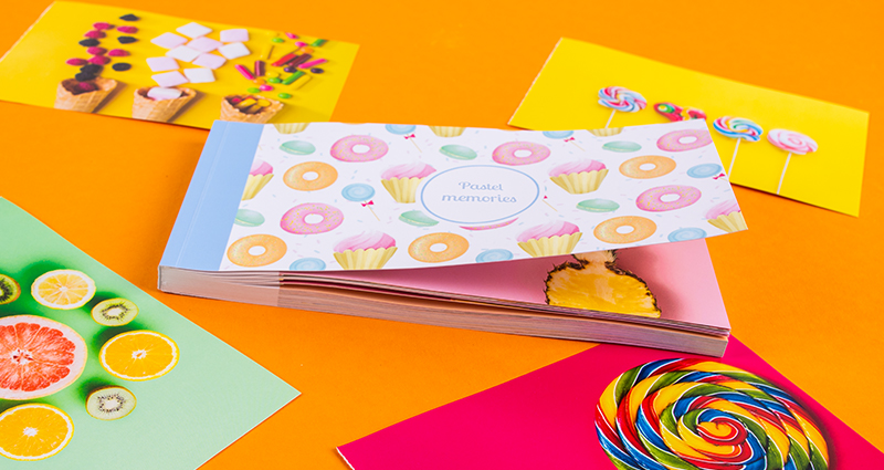Sharebookas su pakeltu viršeliu Pasteliniai Saldainiai, aplink išsidėstytos išplėštos nuotraukos su ryškių spalvų saldainiais.