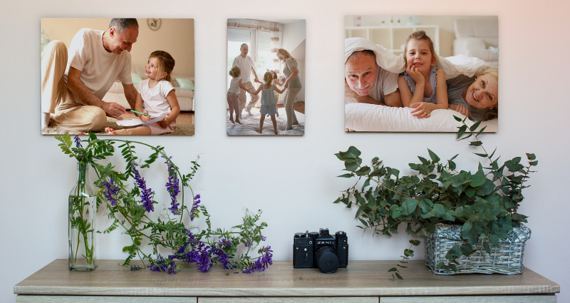 Trois toiles avec les photos de famille accrochées sur un mur claire audessus d’un meuble gris. Sur le meuble deux flacons  avec des fleurs et  un appareil photo .
