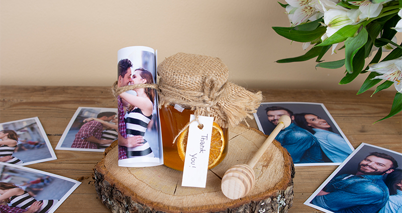 A jar of honey decorated with jute and a rolled up print on the wooden disc, a honey dipper and prints of a couple next to it and white flowers bouquet in the background. 