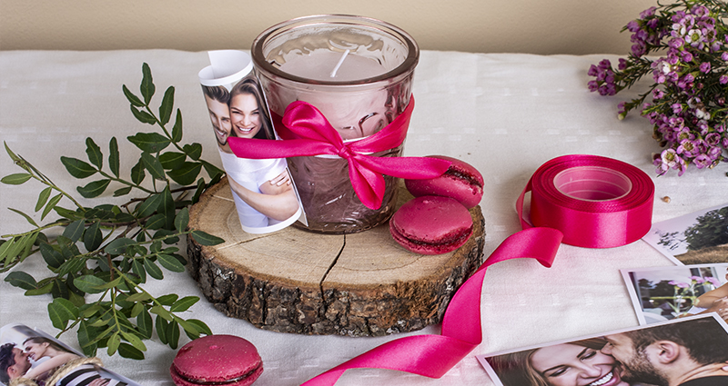 A pink candle in a jar with a rolled-up picture of a couple tied with pink ribbon on a wooden disc. Pink macarons, prints, spool of ribbon, green decorative brunch and tiny pink flowers placed around. A composition on a white tablecloth.