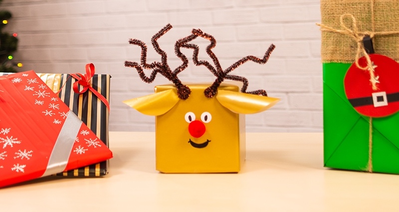 2 livres emballés, une boîte en forme de rennne et un cadeau emballé en style kimono sur un bureau. Au fond un mur de  brique, le texte (Merry) Christmas et un arbre de Noël.