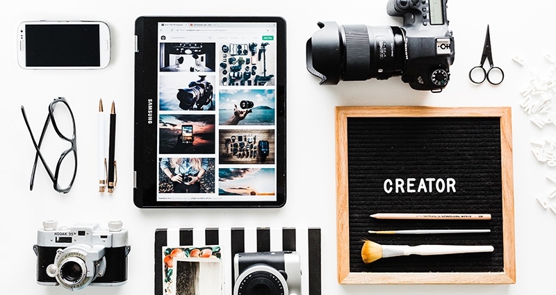 Kameras, ein Smartphone, ein Tablet und andere Gegenstände auf einem weißen Schreibtisch, daneben eine Tafel mit Holzrahmen mit dem Wort "Creator". 