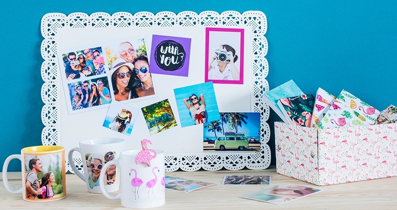 Collection of summer photo products lying on the desk – photo magnets on a white board, sharebooks in a box, photo mugs (colored and latte) and photo prints