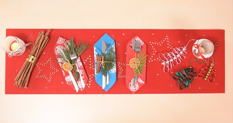 "DIY padaryk pats" Kalėdiniai papuošalai: dekoratyvinės servetėlės skirtos virtuvės įrankiams, žibintai iš stiklainių ir kalėdinės eglutės, pagamintos iš juostelių, išdėstytos ant raudonos servetėlės, kairėje ilgoji cinamono lazdelė.