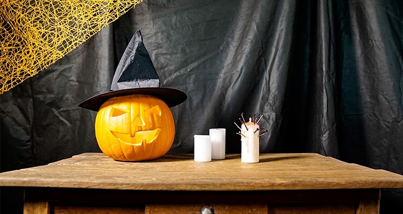 Una calabaza de Halloween con un sombrero de bruja puesto. Al lado tres velas, todo esto puesto en un escritorio marrón viejo. En el fondo una cortina negra con una red naranja en la esquina.