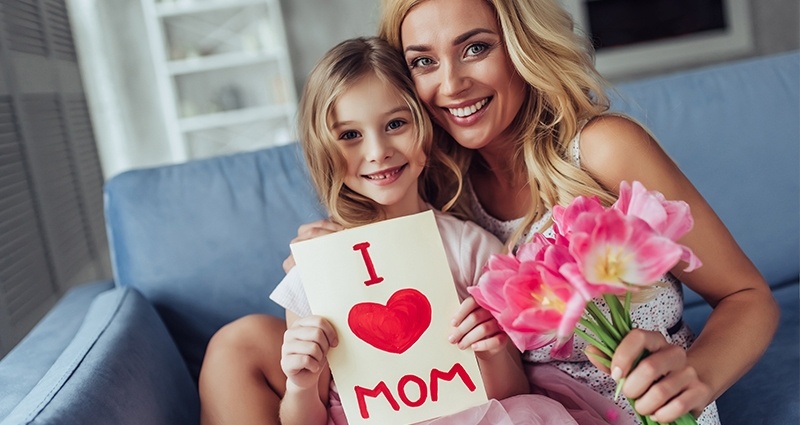 Mom and daughter are sitting on a grey couch. Mom is holding a bouquet of flowers and the girl a Mother’s Day card with the “I <3 MOM” caption.