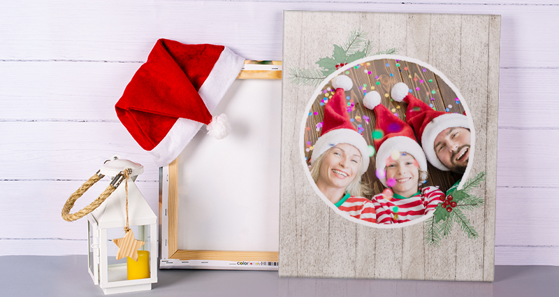 Fotolienzo de Navidad, detrás otro fotolienzo volteado con un gorro de Santa Claus. Al lado un farol blanco. En el fondo una pared de madera.