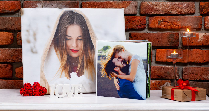 Sur une commode blanche une grande toile photo et un album photo, à coté un cadeau avec le ruban rouge et des cœurs décoratifs et le texte LOVE, au fond le mur en brique rouge