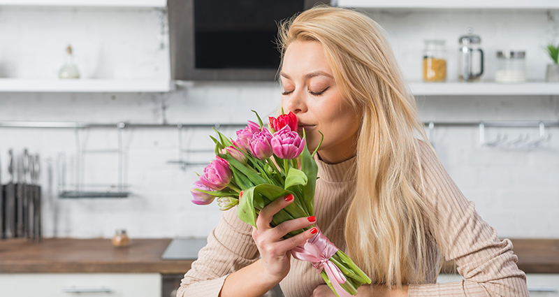 Une femme aux cheveux blondes sentant un bouquet des tulipes à la table de cuisine. Au fond une hotte aspirante et des étagères de cuisine.
