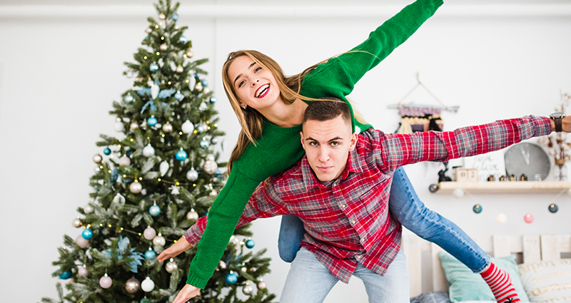 Un garçon qui tient sa copine sur son dos. Au fond un lit et un arbre de Noël  