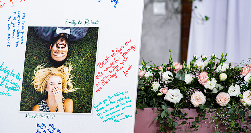 Zoom su una grande tela con al centro la foto di una coppia di innamorati e intorno le dediche lasciate dagli invitati con dei pennarelli colorati. Sullo sfondo c'è un bouquet di fiori in colori pastello su un tavolo decorato con un tulle rosa cipria.