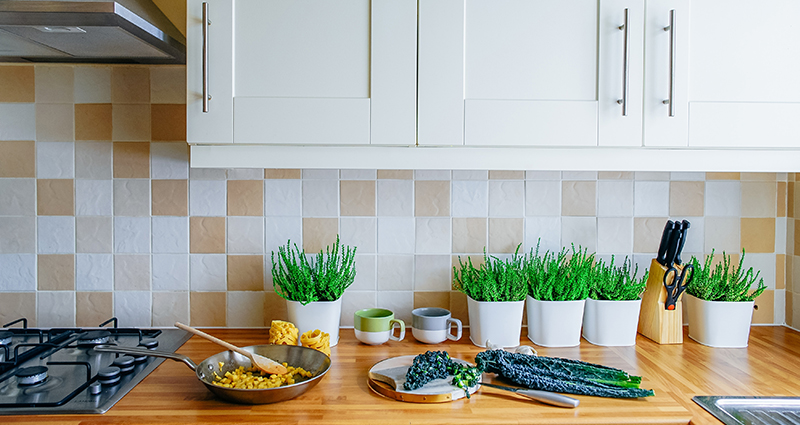 El zoom sobre una encimera clara, sobre la cual hay hierbas aromáticas en macetas blancas y  verduras llenas de color, más arriba un armario blanco.