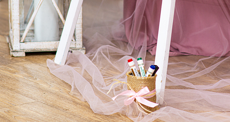 A close-up on a can with colourful marker pens. The can is decorated with a hemp string and a pink ribbon. It is standing on the floor on pink netting placed around a white easel. In the background there’s a table and a white lantern